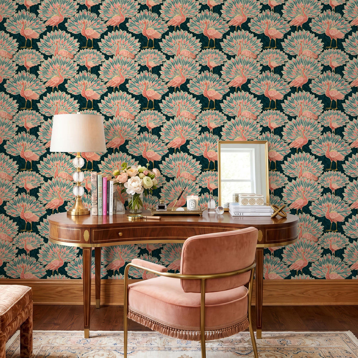 May include: A curved wooden desk with a lamp, a vase of flowers, and a mirror, positioned against wallpaper featuring a repeating pattern of pink peacocks on a dark teal background. A pink upholstered chair with fringe sits in front of the desk.